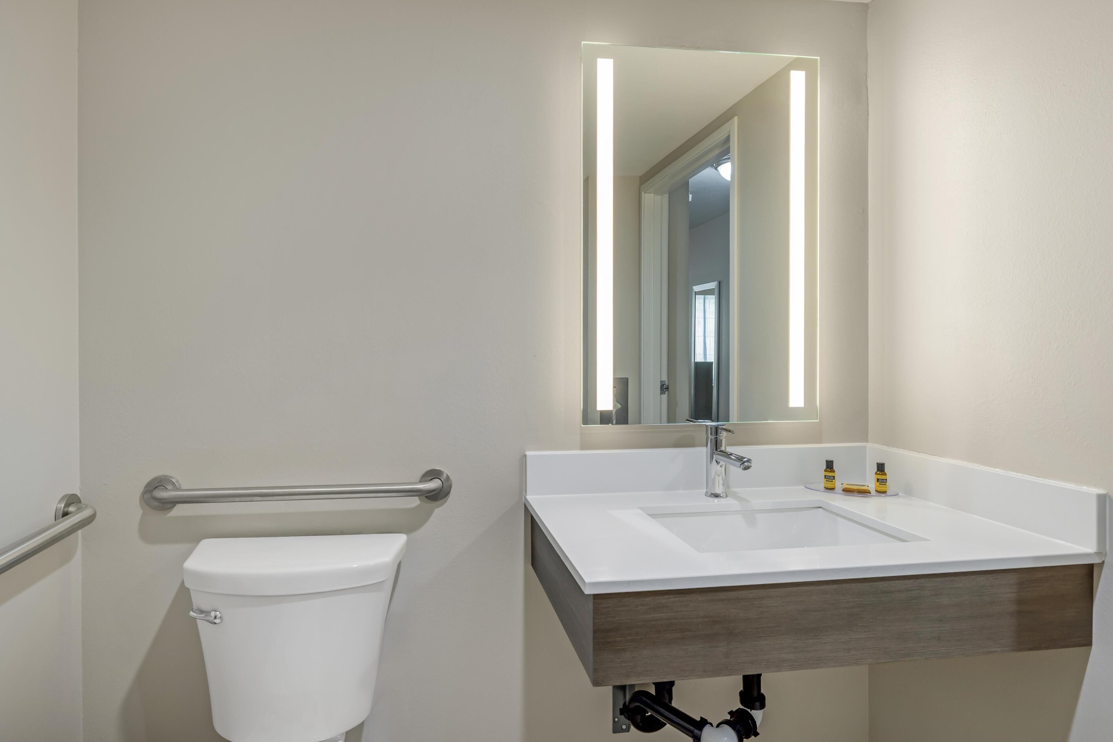Une salle de bain avec des toilettes blanches, un lavabo surmonté d’un miroir et une barre d’appui.