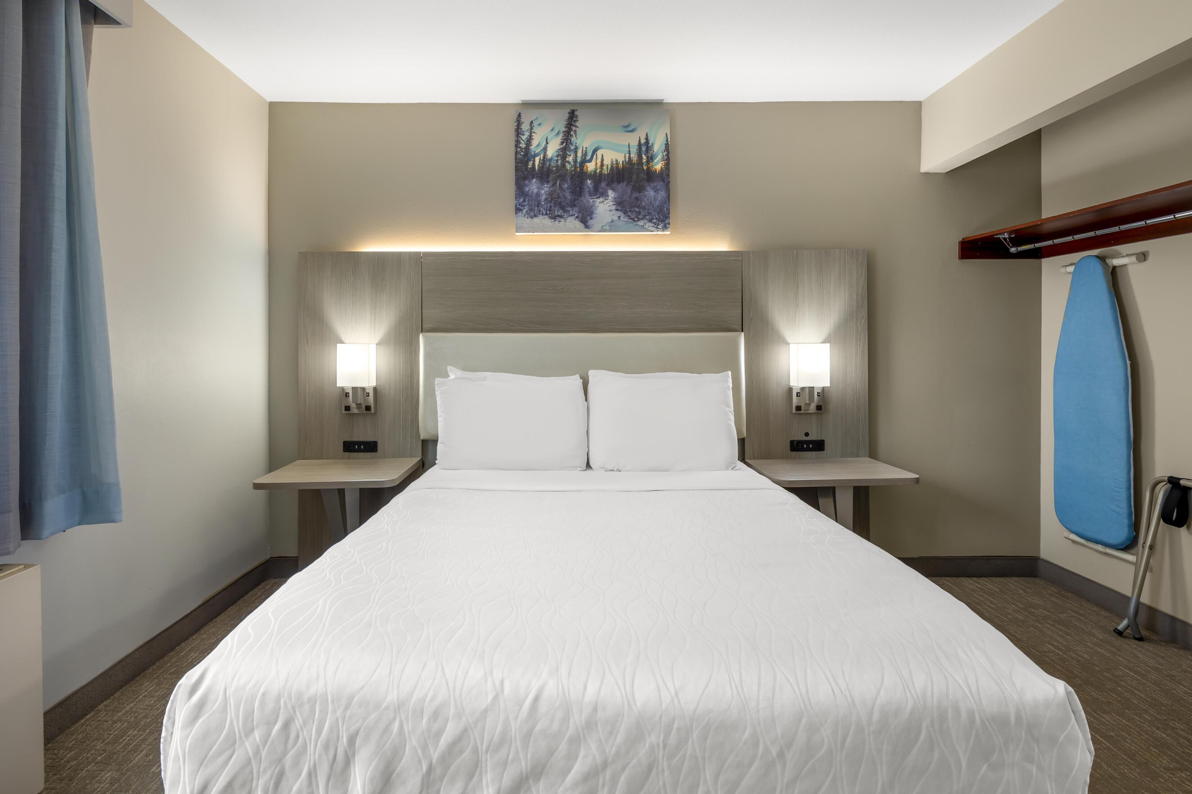 A hotel room with a large bed, two bedside tables with lamps, a painting on the wall, and a blue ironing board.