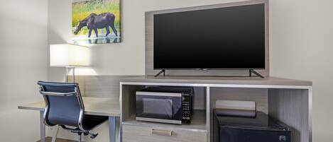 A hotel room with a desk, a flat-screen TV, a microwave, a refrigerator, a chair, and a painting on the wall.