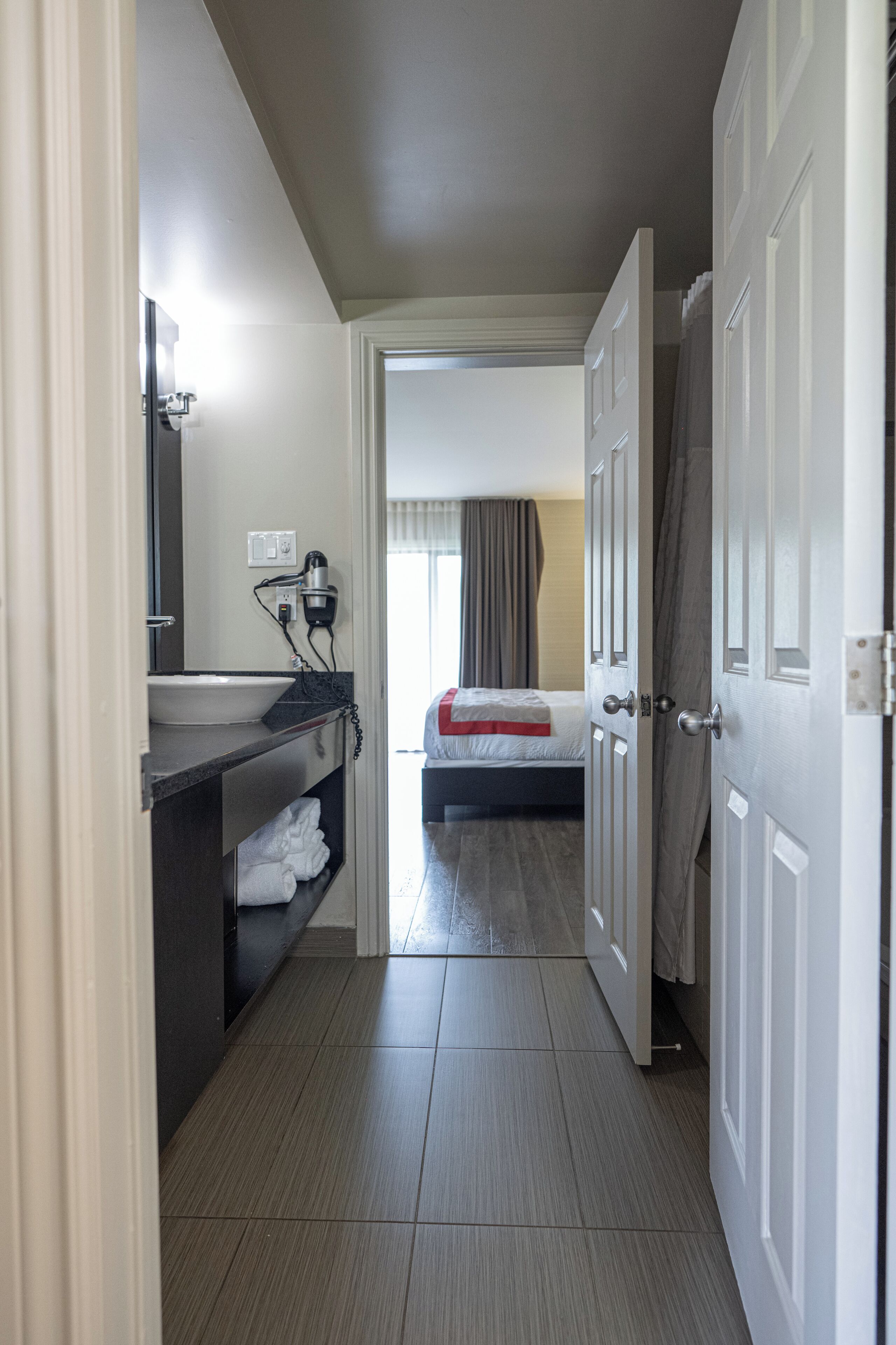 A narrow hallway with a bed, a bathroom with a sink and mirror, and a door leading to another room.