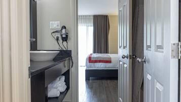 A narrow hallway with a bed, a bathroom with a sink and mirror, and a door leading to another room.