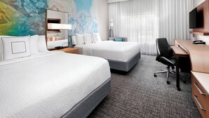 A hotel room with two beds, a desk, a TV, and a bathroom with a mirror and sink.