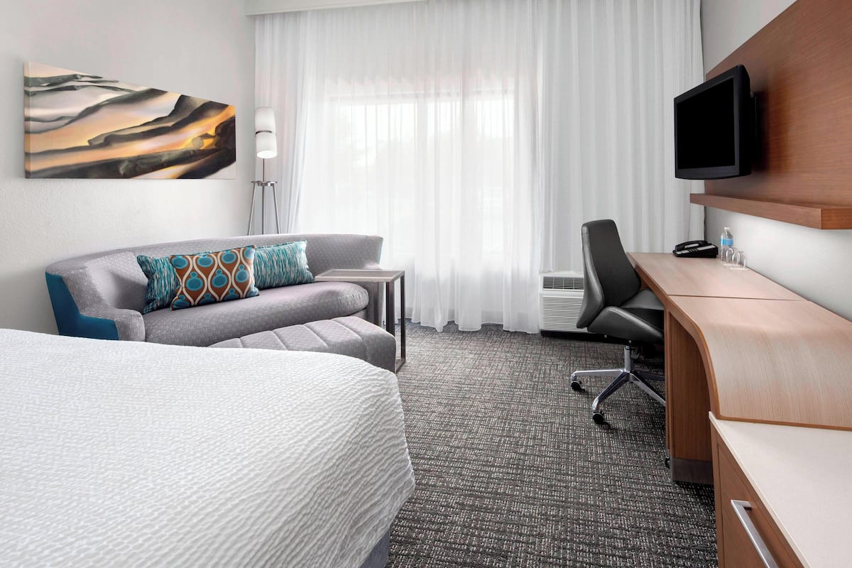 Una habitación de hotel con cama, sofá, escritorio, televisión y un amplio ventanal con cortinas.