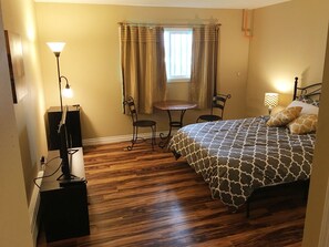 A bedroom with a wooden floor, a bed with patterned bedding, a desk with a lamp, a chair, and a window with curtains. - Travel Inn (Anchorage)