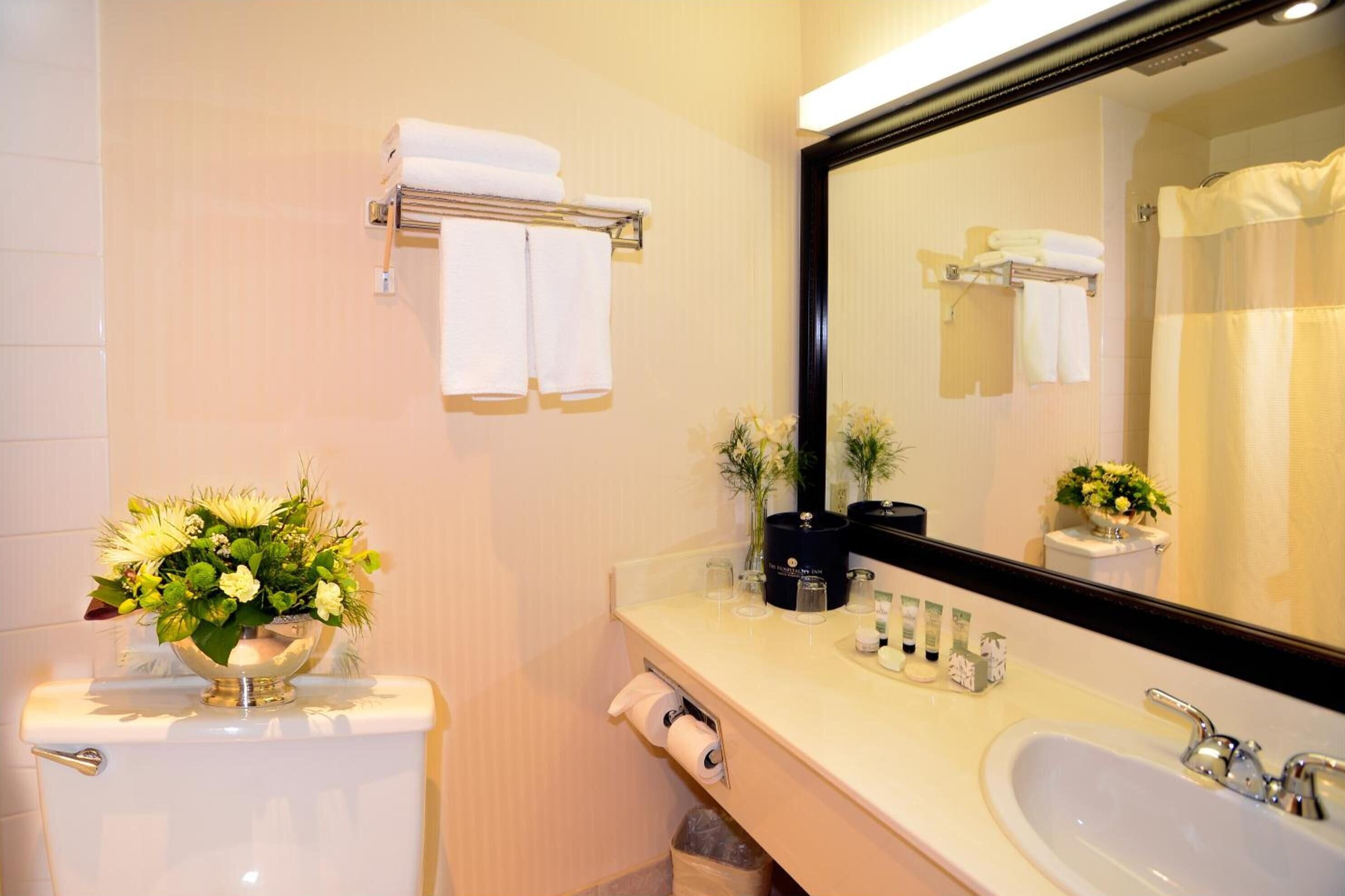 Un baño con inodoro, lavamanos y espejo. Hay toallas en un perchero y un arreglo floral sobre el inodoro.