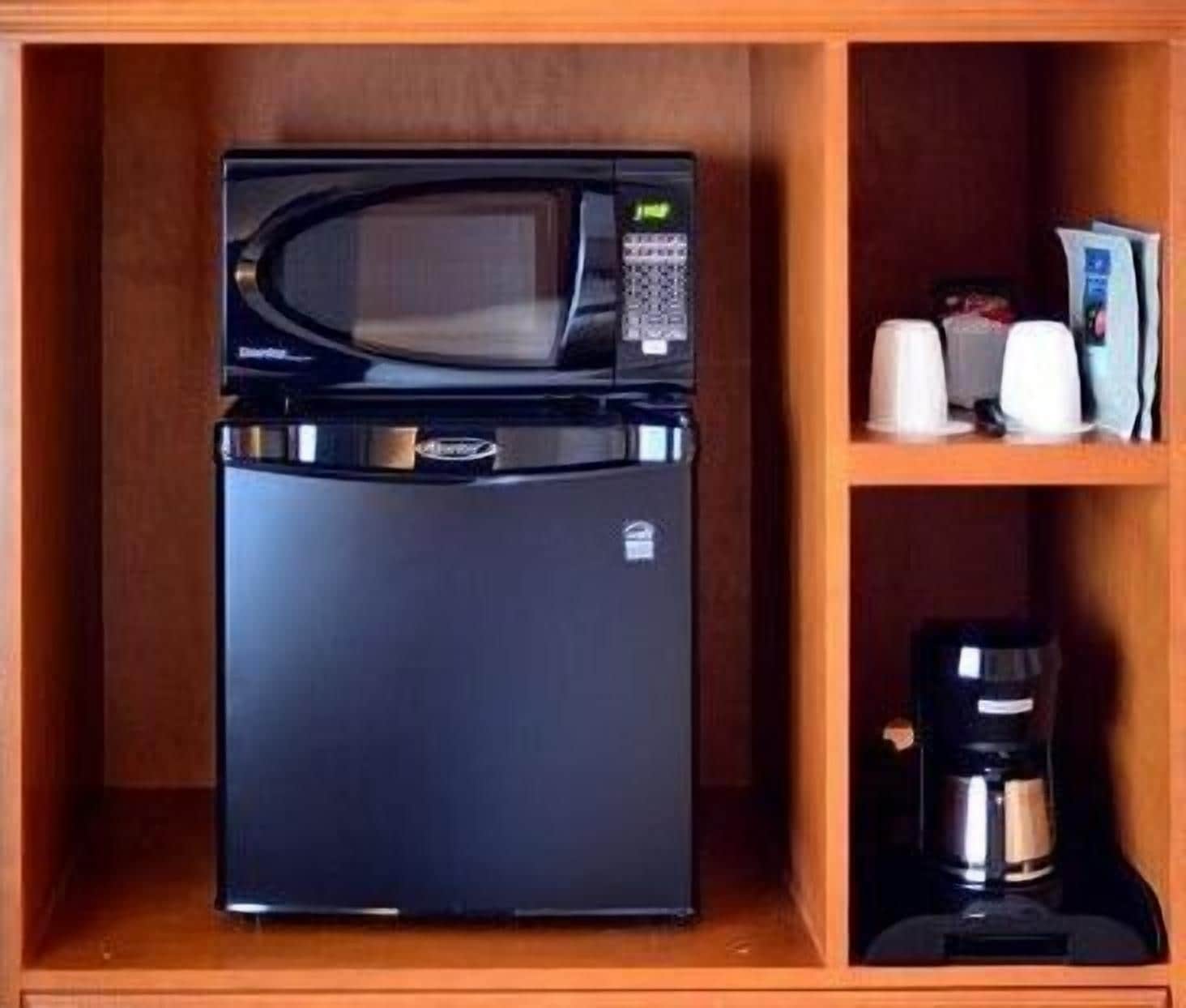 Un refrigerador negro con un microondas encima, una cafetera y una repisa con tazas y una caja.
