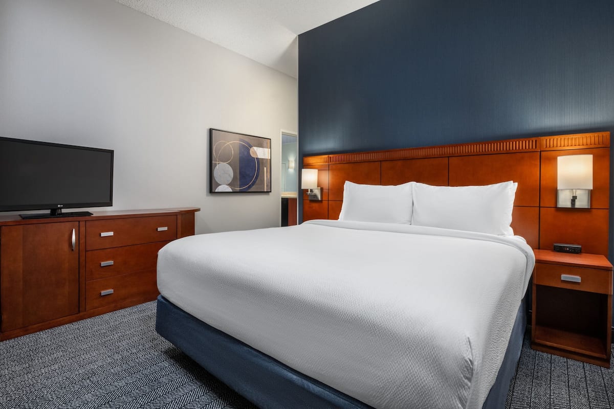A hotel room with a large bed, wooden headboard, bedside lamps, a flat-screen TV, and a dresser.