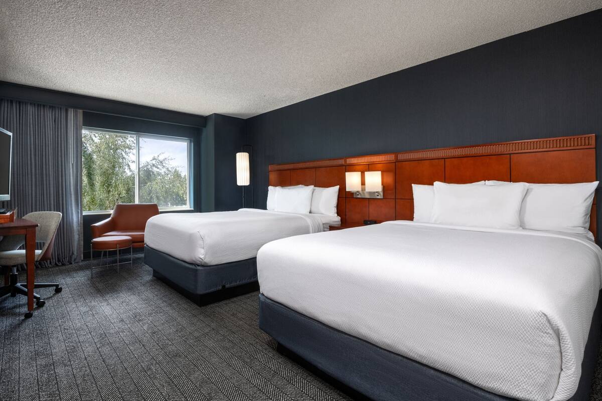 A hotel room with two beds, a desk, a chair, and a large window.
