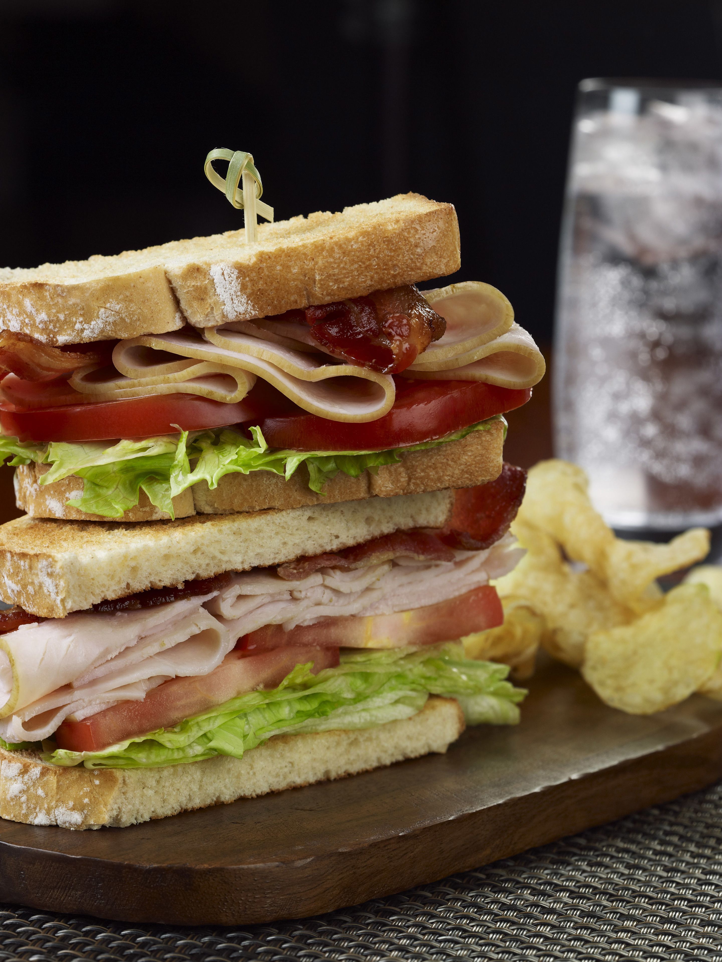 A sandwich with multiple layers of meat, cheese, and vegetables, accompanied by a glass of water and potato chips on a wooden board.