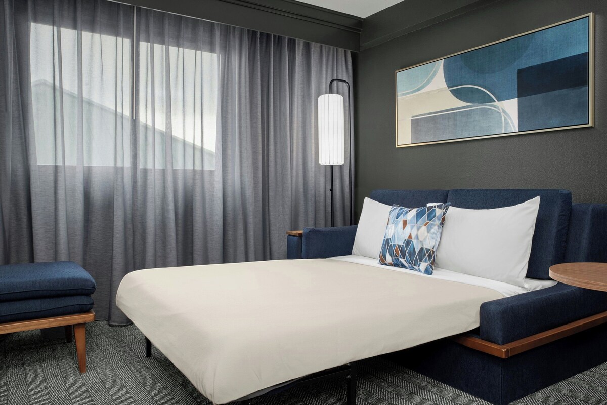 A hotel room with a bed, a blue sofa, a small table, a lamp, and a painting on the wall.