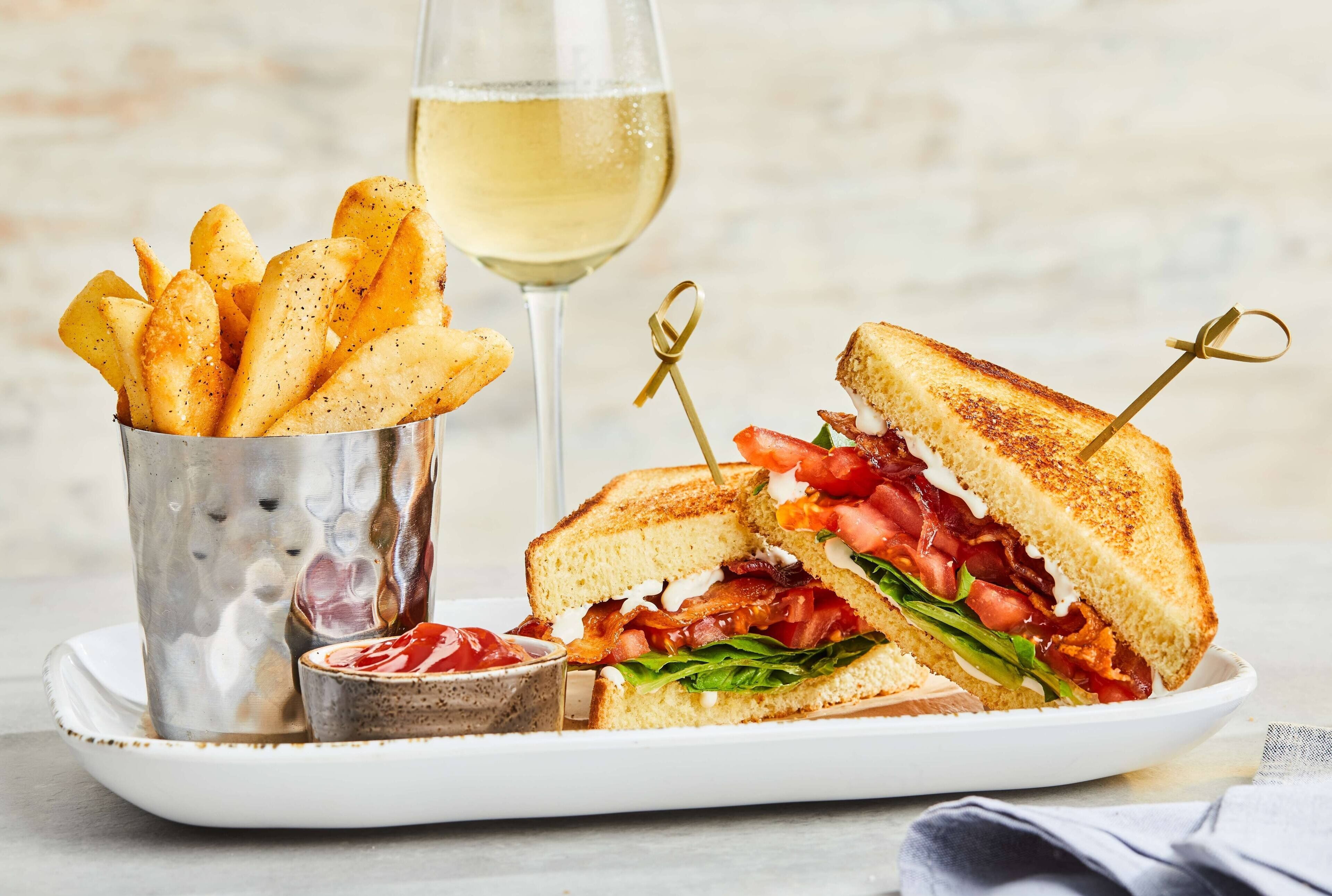 A plate with a grilled sandwich, a glass of white wine, and a container of seasoned fries.