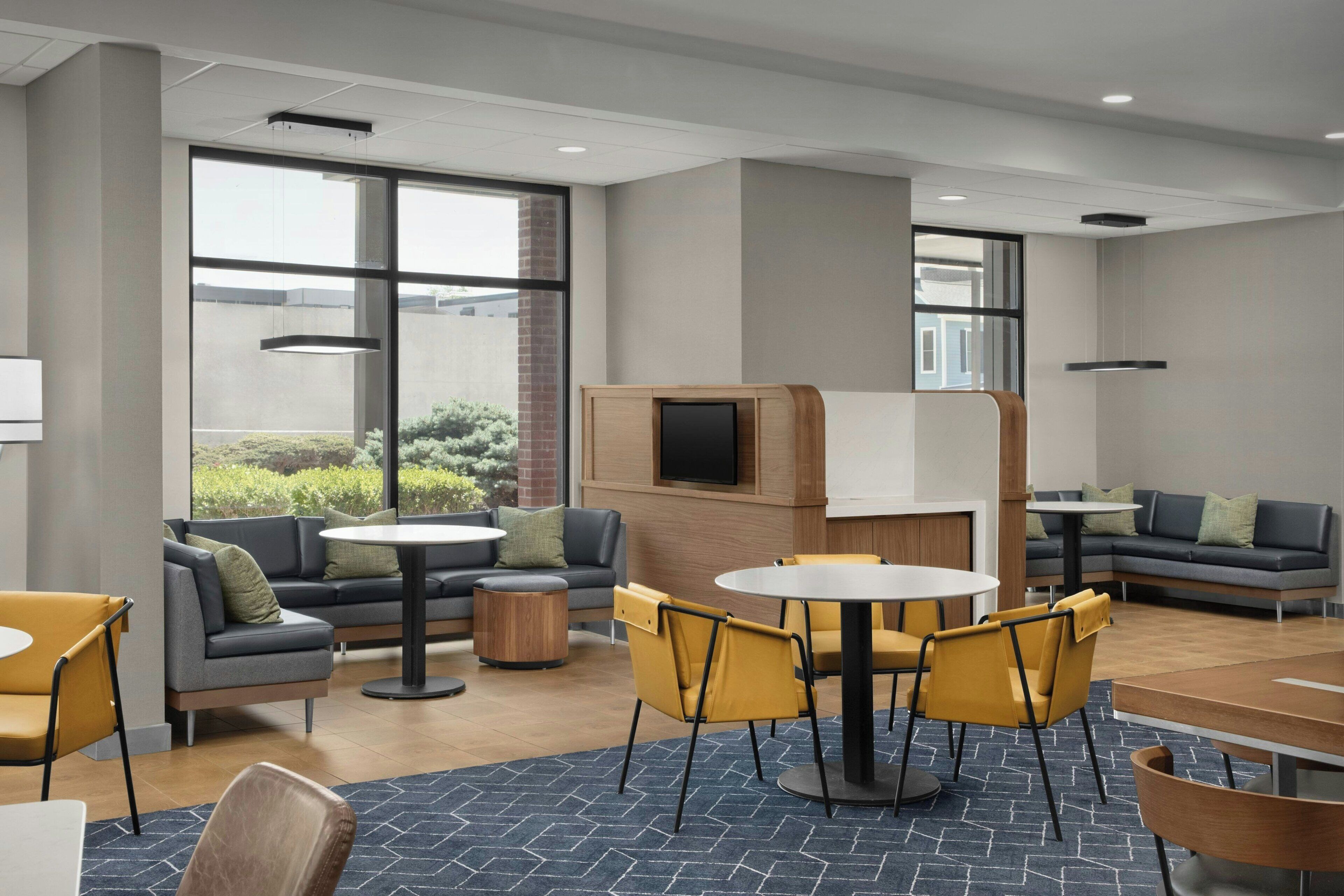 A modern lounge area with sofas, tables, and chairs, featuring large windows and a flat-screen TV.
