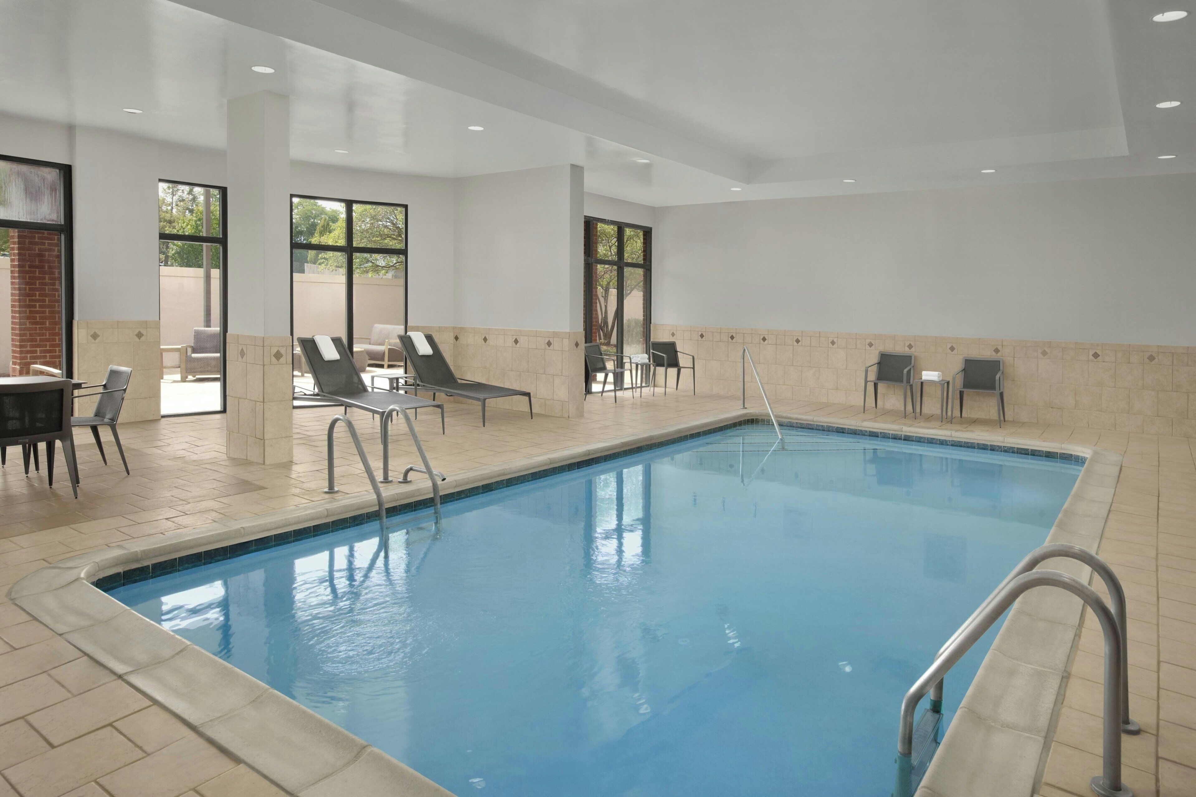 A swimming pool with lounge chairs and a sunbed area, surrounded by tiled walls and large windows.