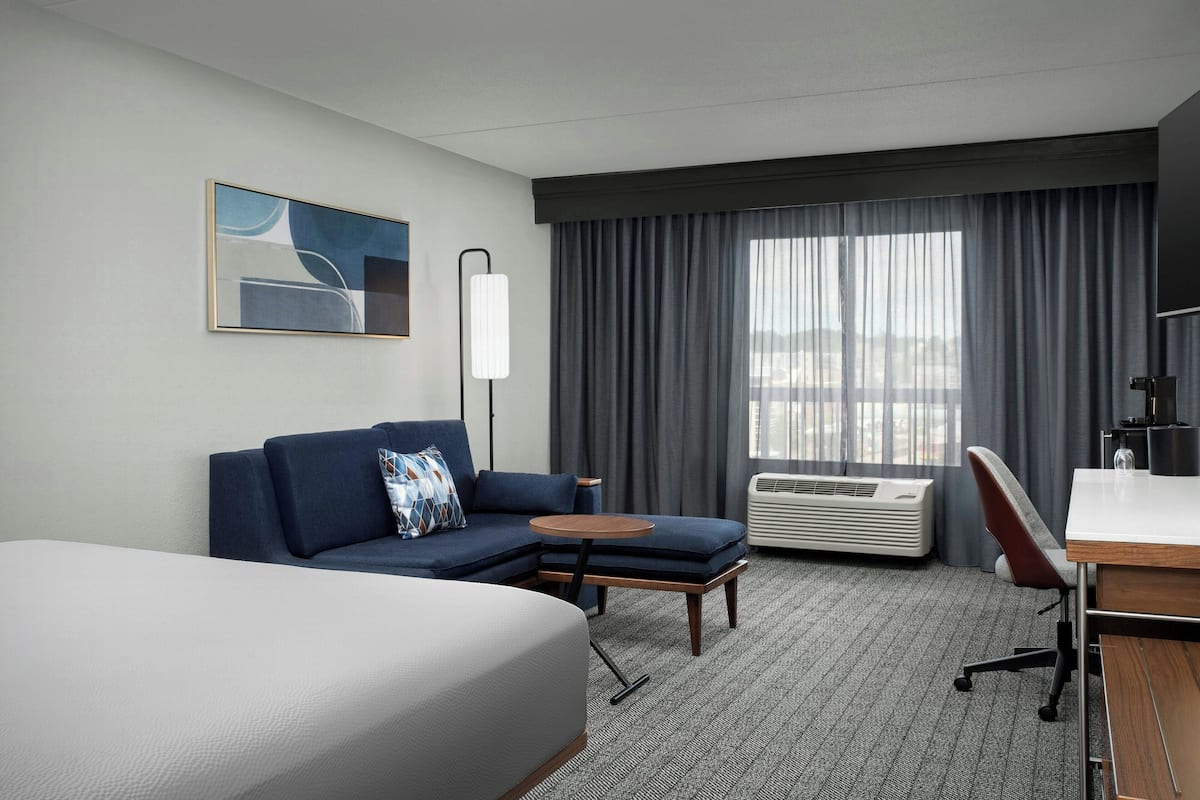 Une chambre d’hôtel avec un lit, un canapé, une petite table, un bureau et une grande fenêtre avec des rideaux.