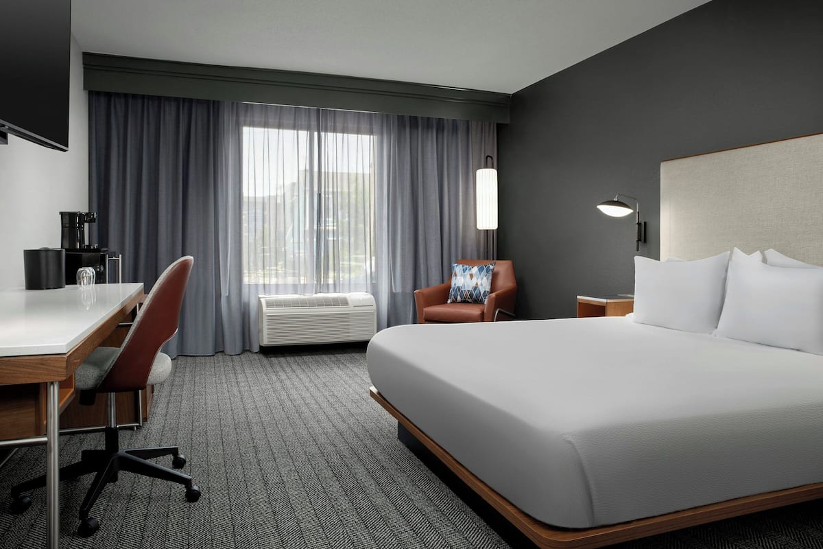 Une chambre d’hôtel avec un grand lit, un bureau avec une chaise, une machine à café et une vue sur la ville par la fenêtre.