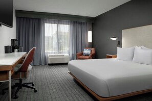 A hotel room with a large bed, a desk with a chair, a coffee maker, and a view of the city through the window. - Courtyard by Marriott Bloomington (Bloomington)