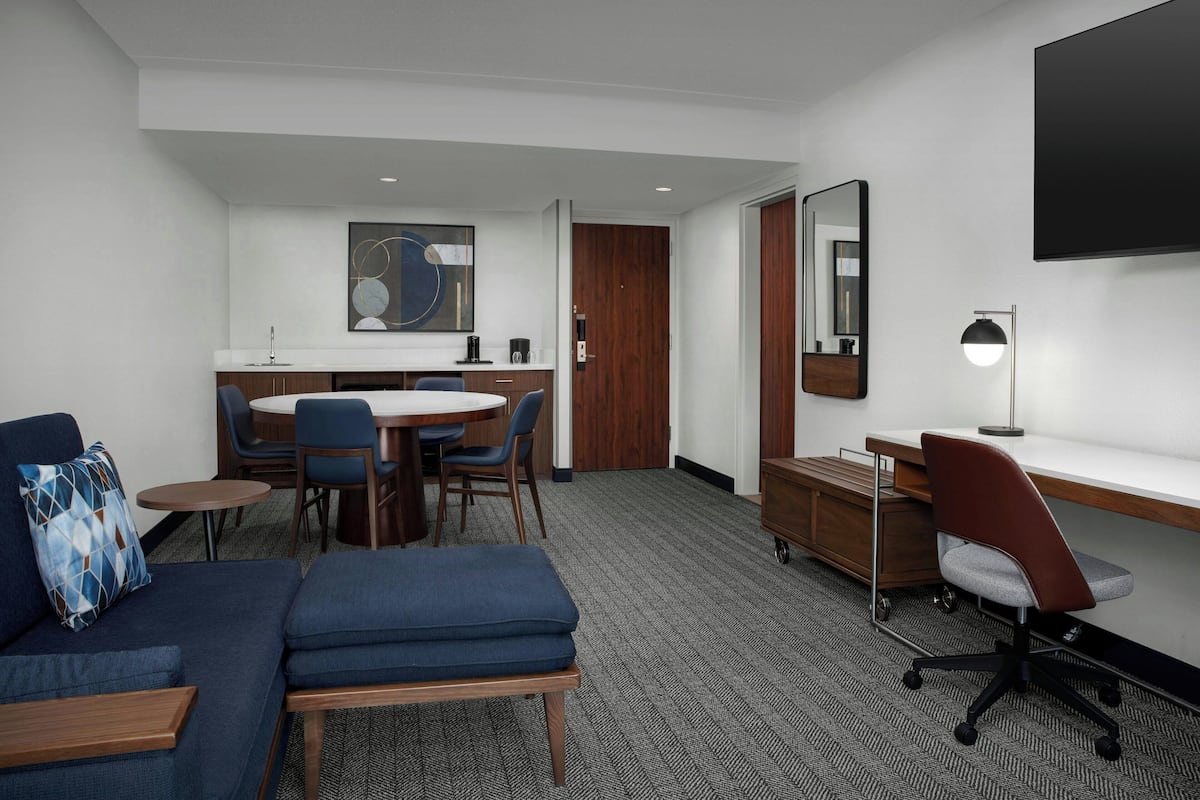 Une chambre d’hôtel comprenant un canapé, une petite table ronde, un bureau avec une chaise, un coin repas avec des chaises, une porte en bois, un miroir et un téléviseur à écran plat fixé au mur.