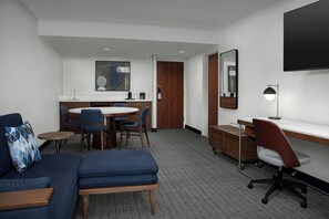 Habitación de hotel con sofá, una pequeña mesa redonda, escritorio con silla, zona de comedor con sillas, puerta de madera, espejo y televisor de pantalla plana montado en la pared.
