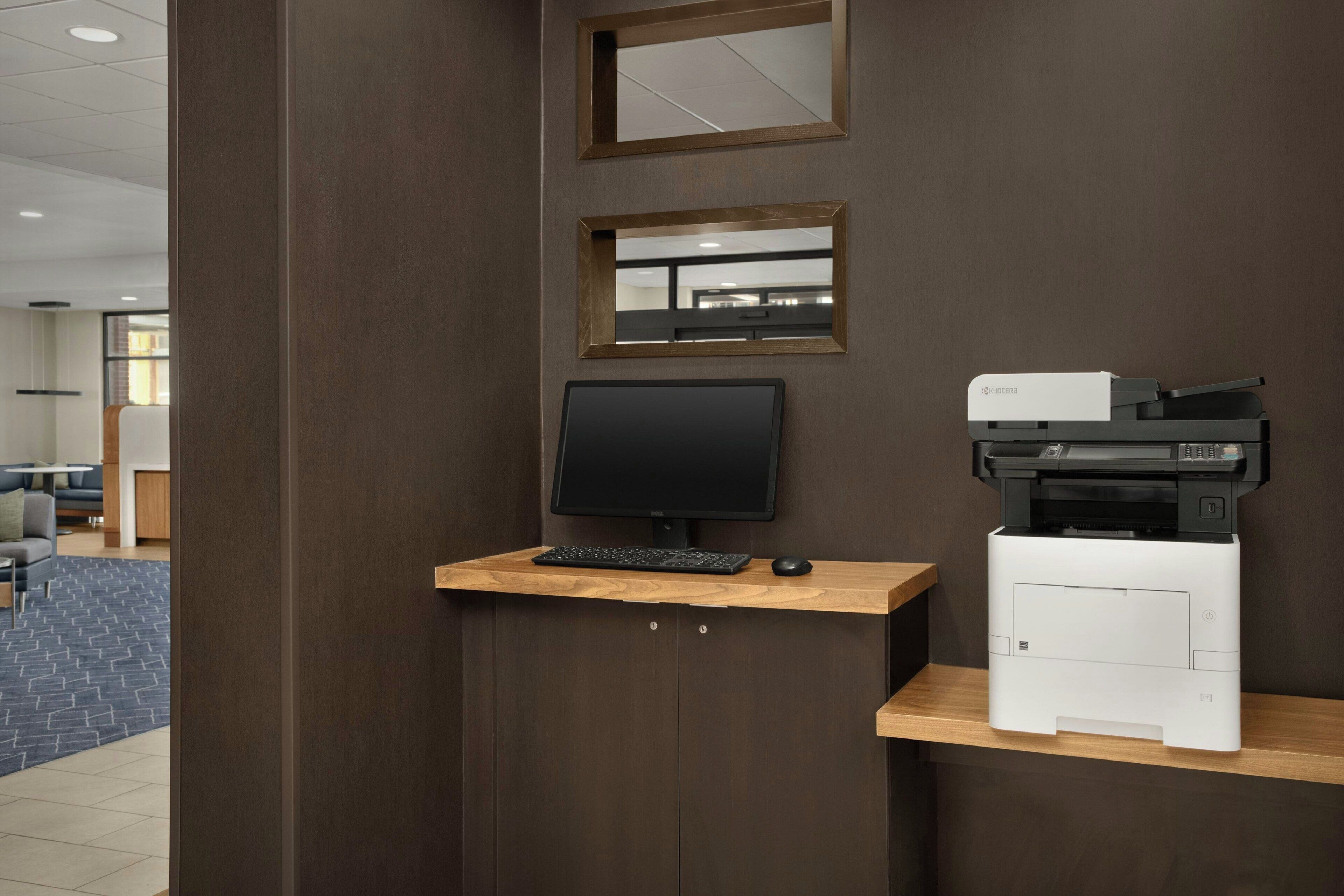 Un espacio de trabajo con monitor de computadora, teclado y una impresora Kyocera sobre un escritorio de madera frente a una pared.