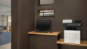 Un espacio de trabajo con monitor de computadora, teclado y una impresora Kyocera sobre un escritorio de madera frente a una pared.