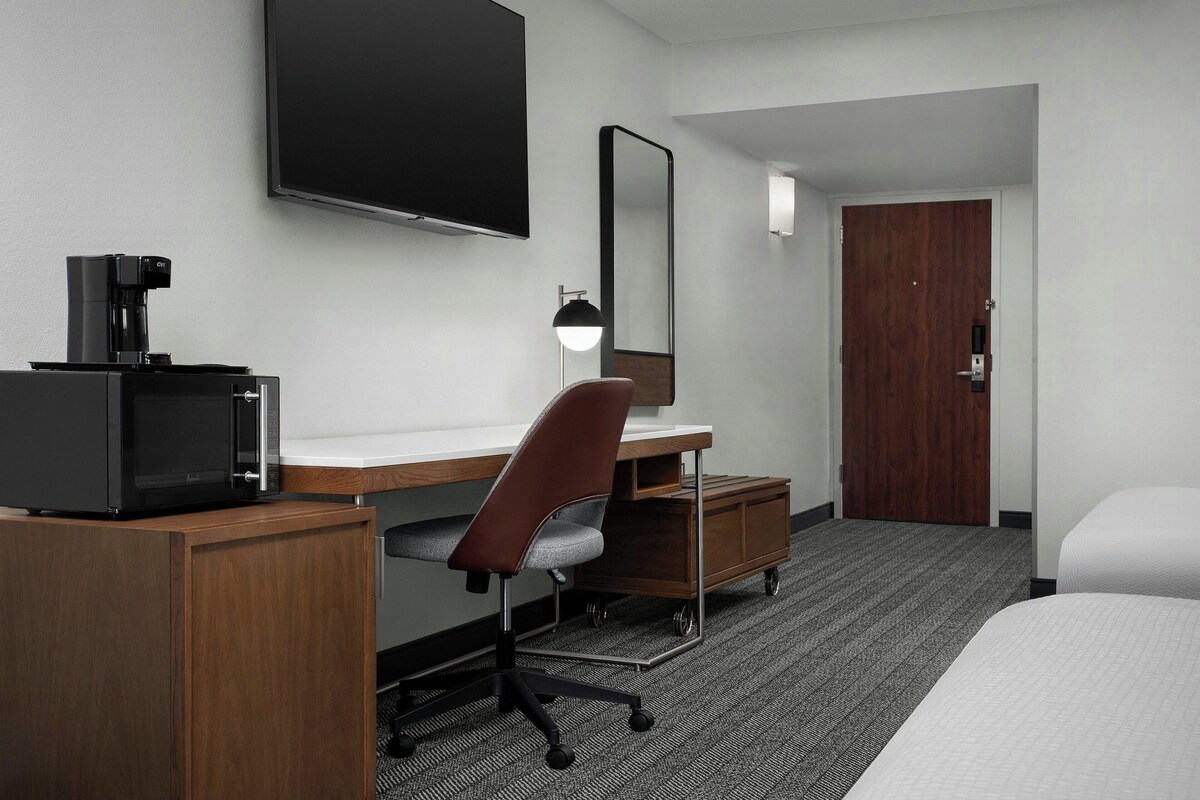 A hotel room with a desk, a chair, a bed, a television, a mirror, and a door.