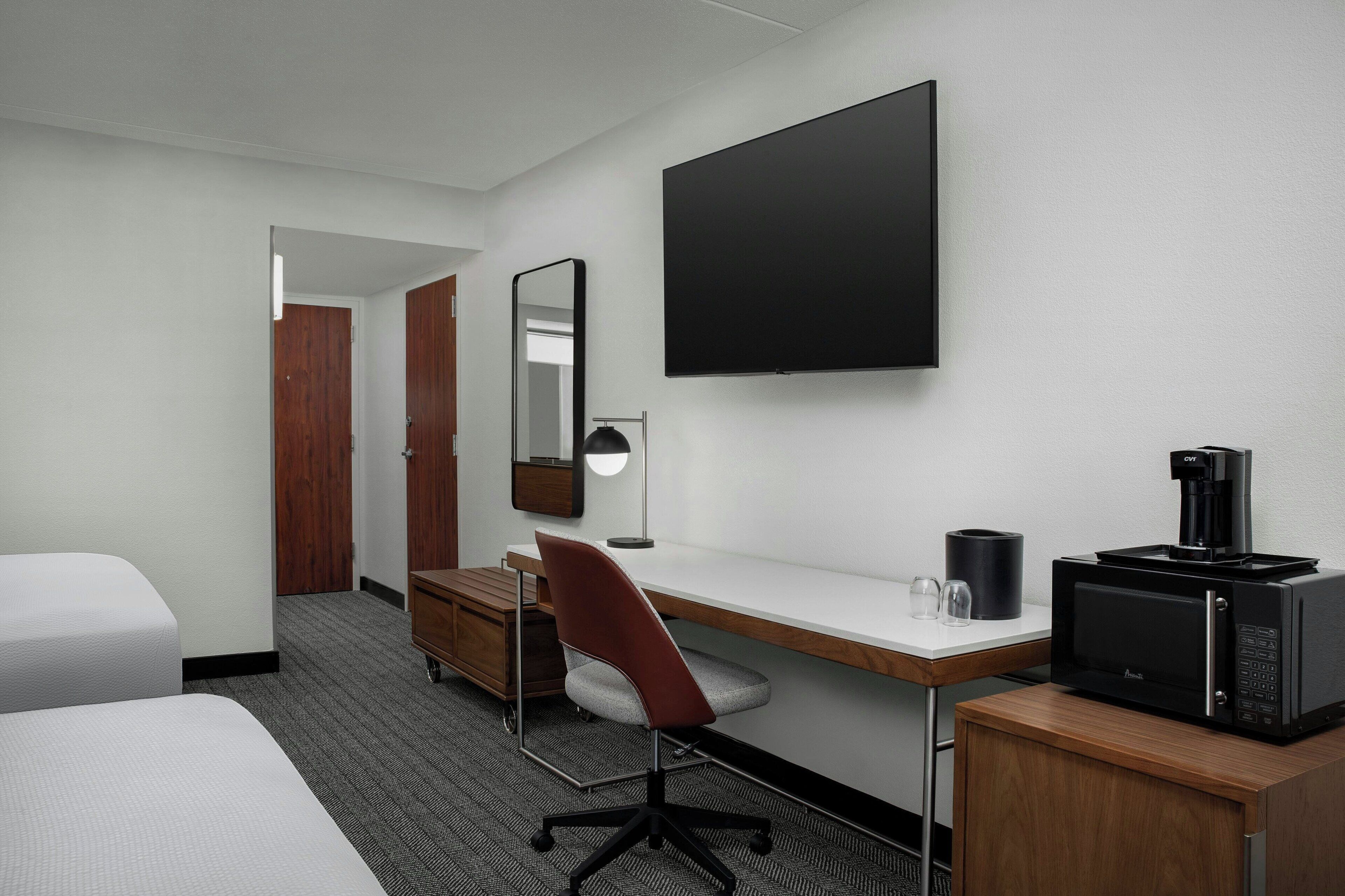A hotel room with a bed, a desk with a computer and a coffee maker, a chair, a mirror, and a flat-screen TV.
