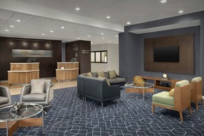 A modern lobby with a dark blue patterned carpet, a grey sofa, a round glass coffee table, and a flat-screen TV mounted on the wall.