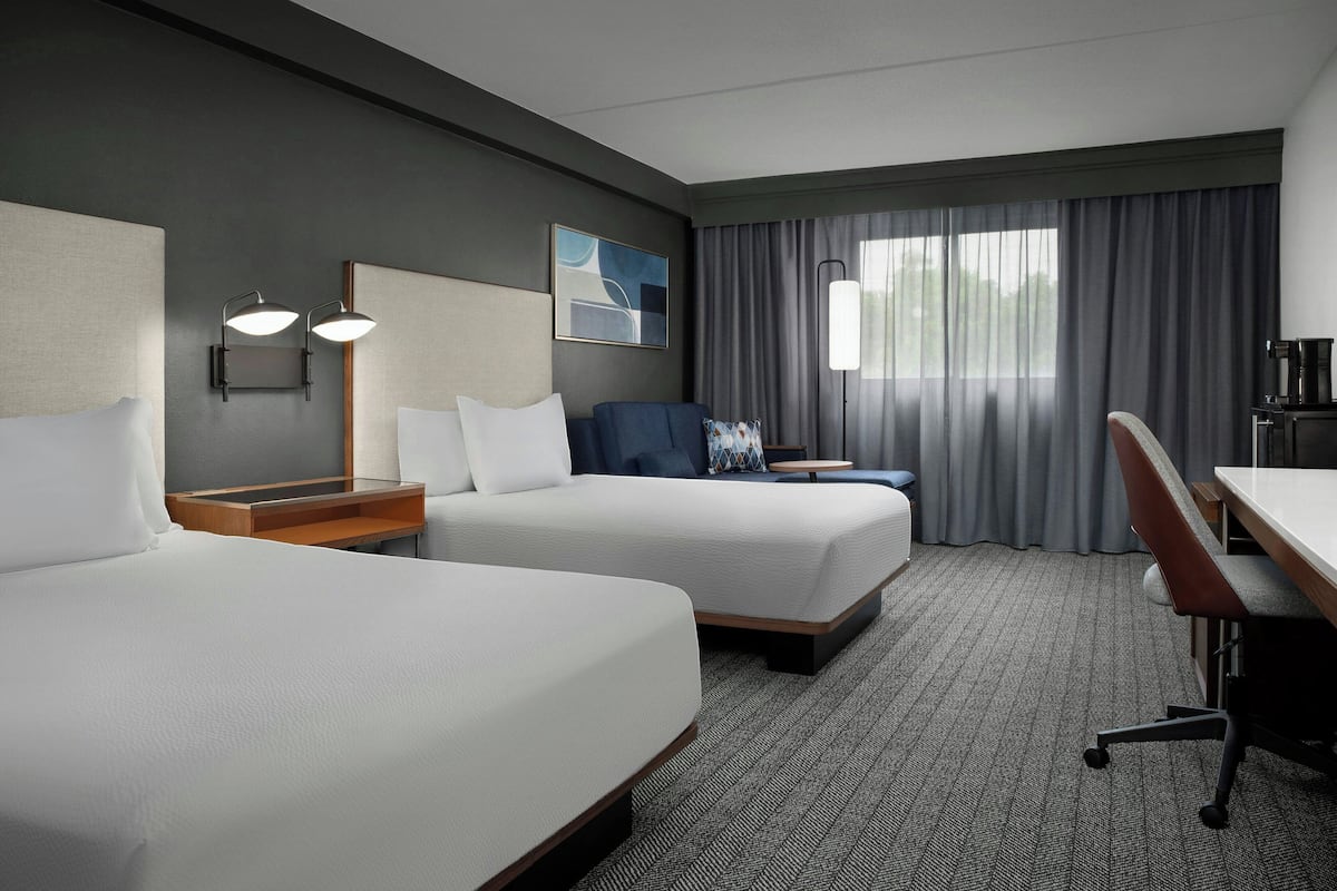 Une chambre d’hôtel avec deux lits, un bureau, une chaise, une lampe et une fenêtre avec des rideaux.