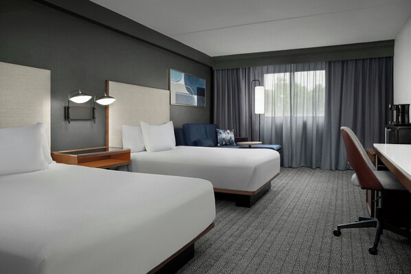 A hotel room with two beds, a desk, a chair, a lamp, and a window with curtains. - Courtyard by Marriott Bloomington (Bloomington)