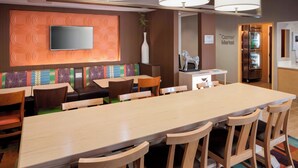 A modern hotel lobby with a long dining table, comfortable seating, a flat-screen TV, a refrigerator, and a sign indicating the 'Corner Market'. - Fairfield Inn Tuscaloosa by Marriott (Tuscaloosa)
