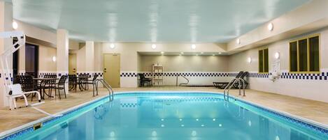 An indoor swimming pool with a tiled border, seating area with chairs, and a changing room in the background.