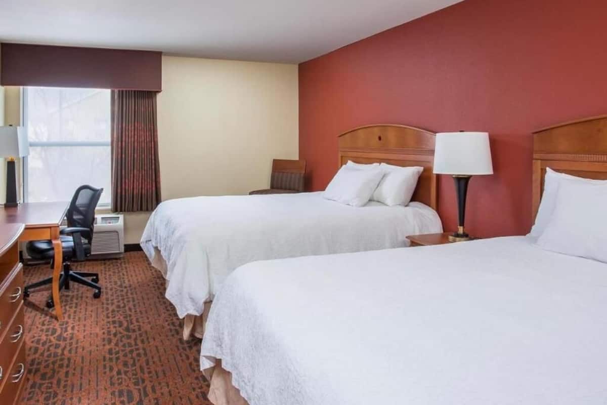 Quarto de hotel com duas camas, uma escrivaninha, uma cadeira, um abajur e uma janela com cortinas.
