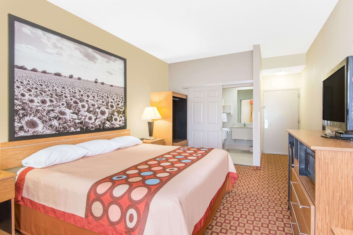 A hotel room with a bed, a large framed picture, a TV, a desk, and a bathroom visible through an open door.
