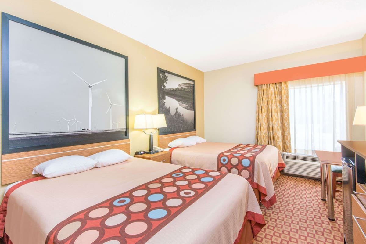 A hotel room with two beds, a desk, a chair, a lamp, a painting of wind turbines, and a window with curtains.