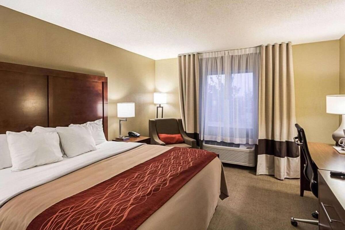 A hotel room with a large bed, a desk, a chair, a window with curtains, and a nightstand with a lamp.