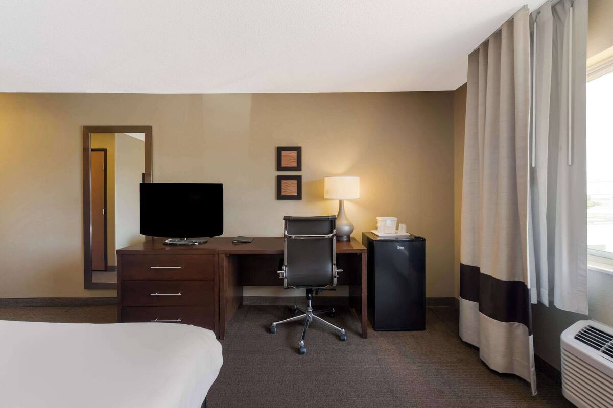 A hotel room with a bed, a desk with a computer, a television, a chair, a lamp, and a mini-fridge.