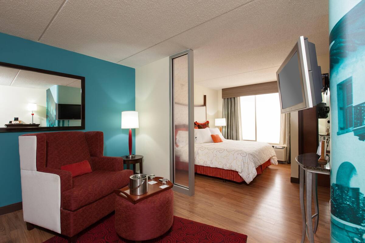 A hotel room with a red armchair, a small table with a tray, a bed with white bedding, a large mirror, and a TV on a stand.