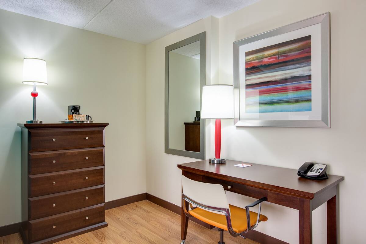A hotel room with a wooden dresser, a desk with a chair, a telephone, a lamp, a mirror, and a framed abstract painting on the wall.