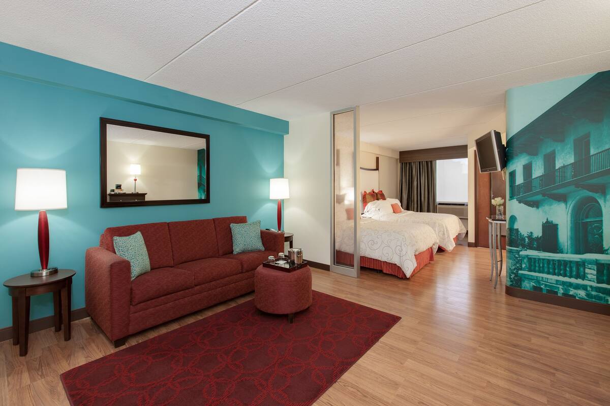 A modern hotel room with a red sofa, a red ottoman, a large mirror, and a painting of a building on the wall.