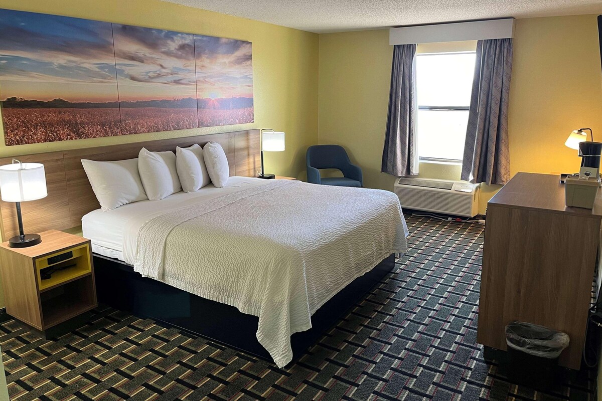 A hotel room with a large bed, a desk, a chair, a wall-mounted TV, and a window with curtains.