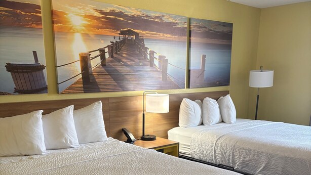 Une chambre d’hôtel avec deux lits, une tête de lit en bois, une table de chevet avec une lampe et un mur orné d’un tableau représentant un coucher de soleil.