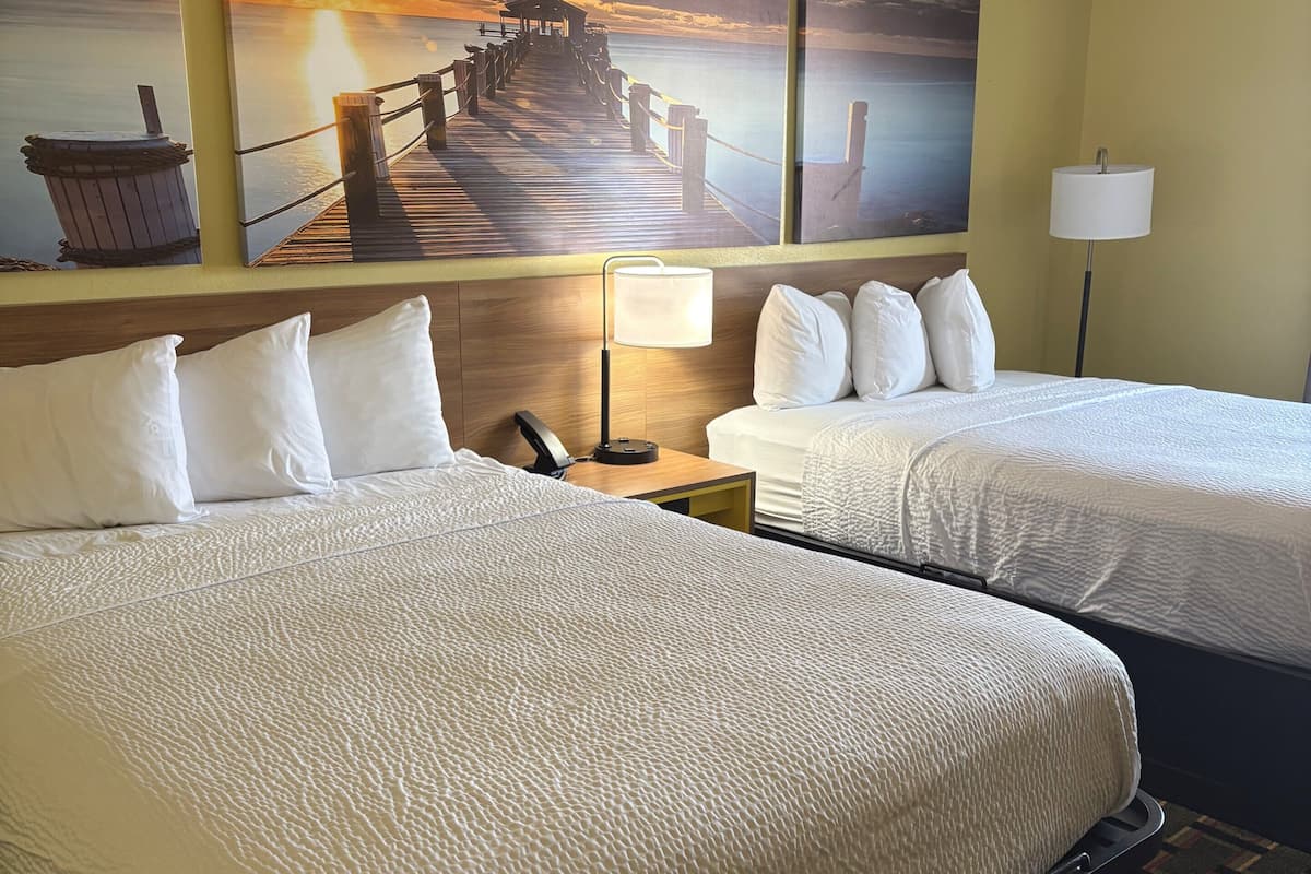 A hotel room with two beds, a wooden headboard, a nightstand with a lamp, and a wall with a sunset painting.