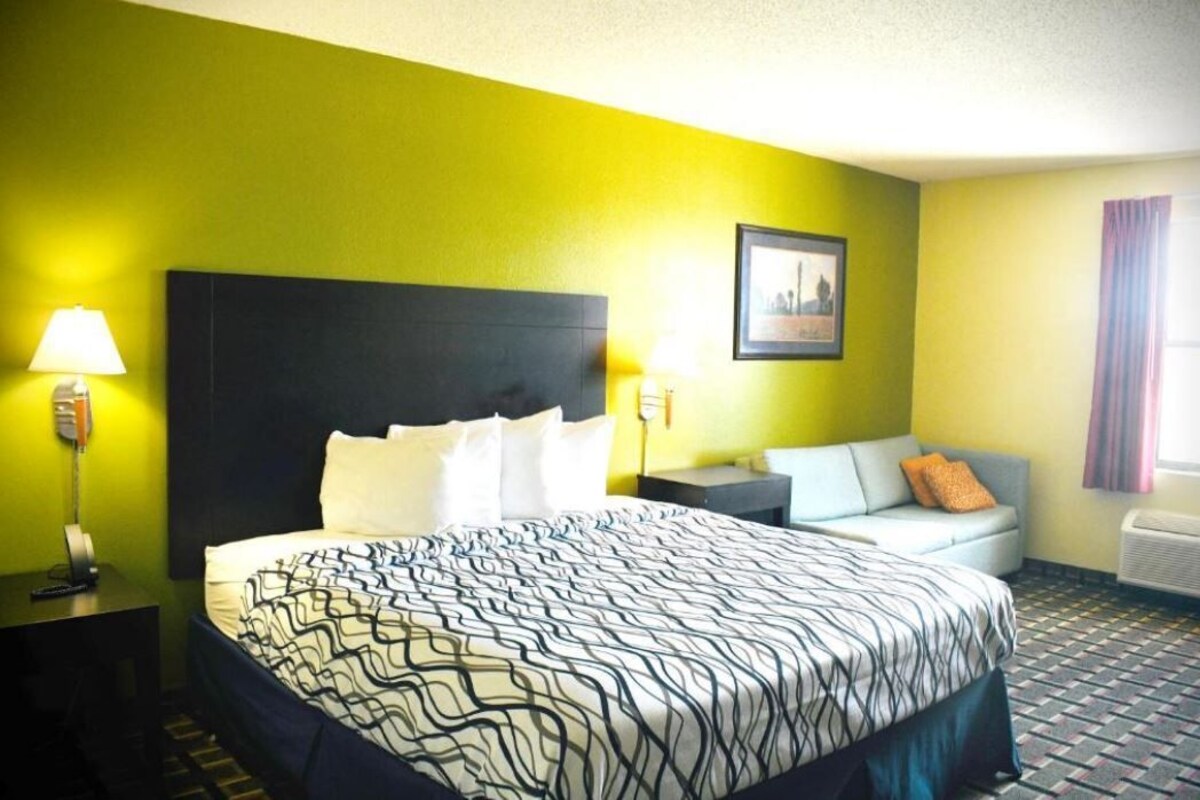 A hotel room with a bed, a nightstand, a lamp, a sofa, and a picture on the wall.