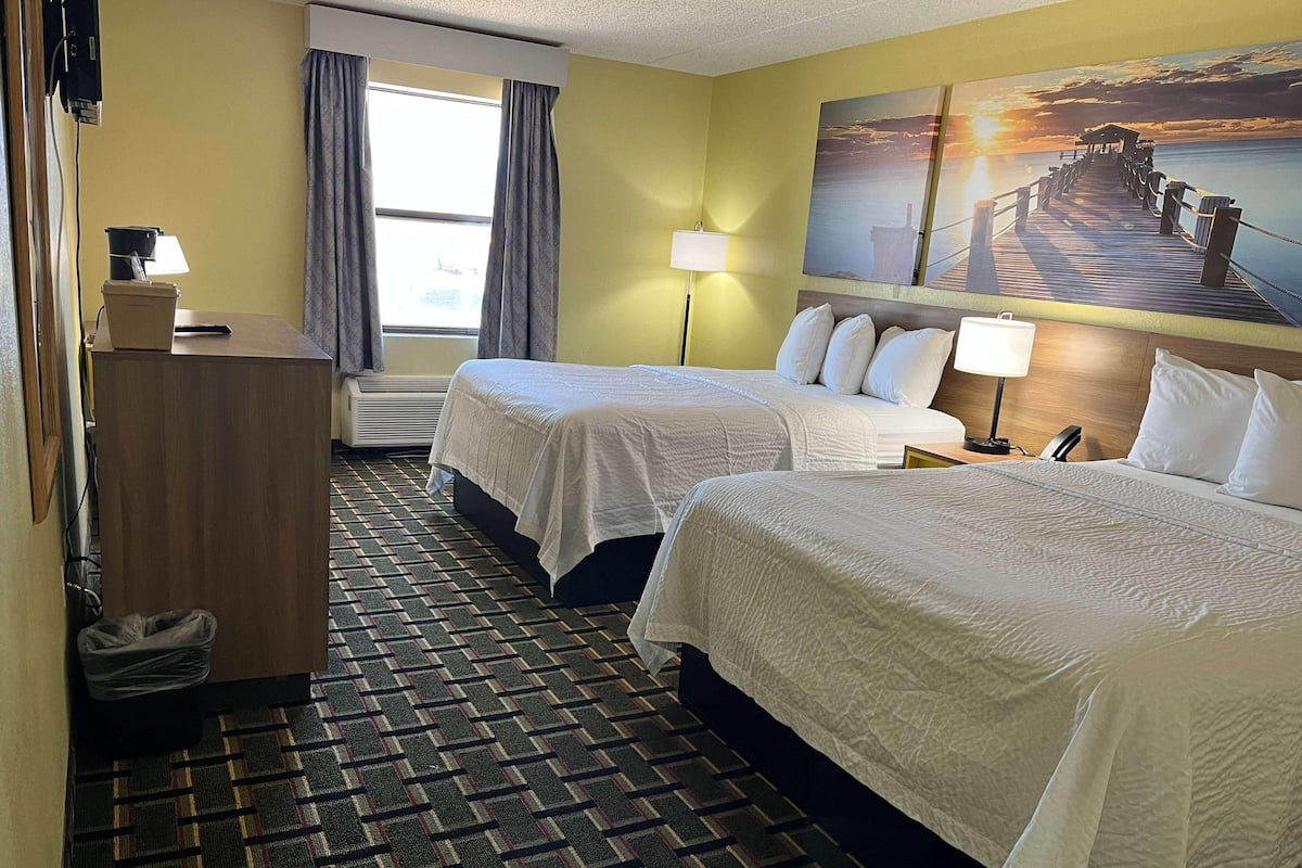 A hotel room with two beds, a desk, a lamp, a painting of a pier, and a window with curtains.