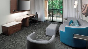 A hotel room with a flat-screen TV mounted on the wall, a desk with a chair, a grey armchair, a blue sofa with a footrest, and a balcony with a view of greenery. - Courtyard Tuscaloosa by Marriott (Tuscaloosa)
