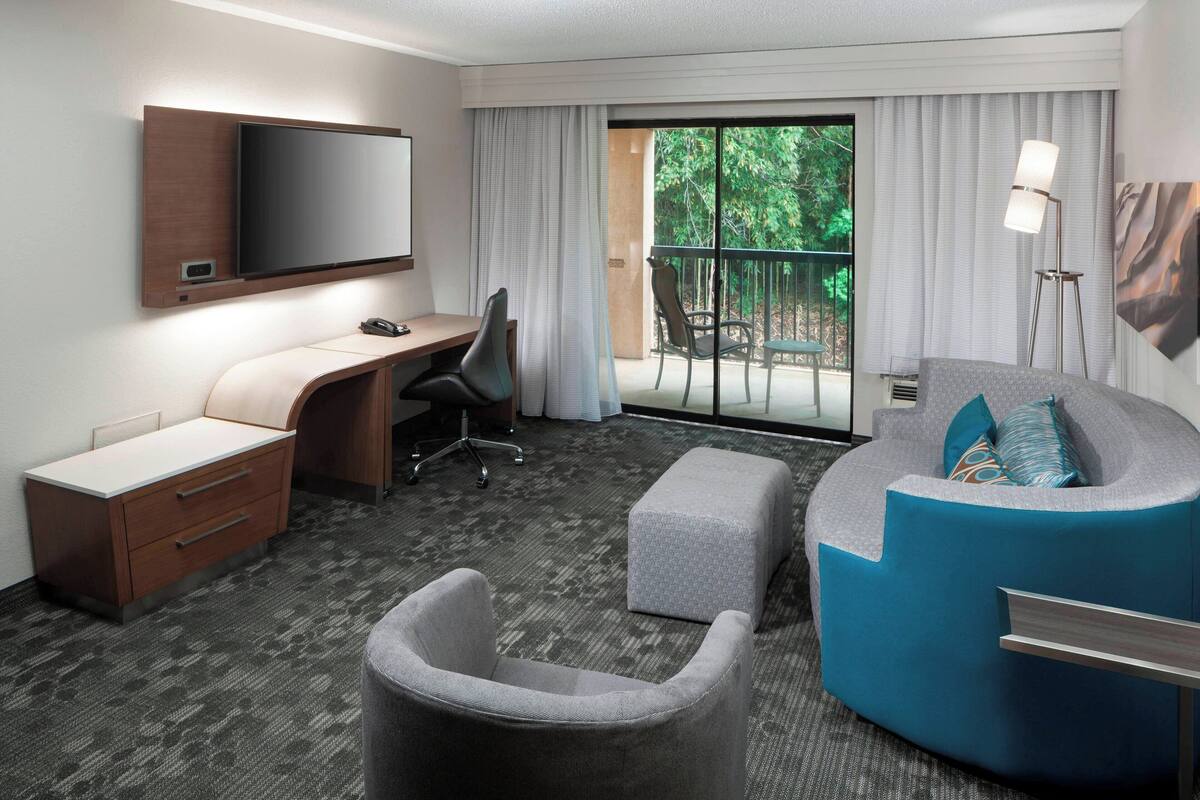 Ein Hotelzimmer mit flachbildschirmfernseher an der Wand, einem Schreibtisch mit Stuhl, einem grauen Sessel, einem blauen Sofa mit Fußhocker und einem Balkon mit Blick auf Grünflächen.