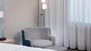 A hotel room with a bed, a grey armchair, a bedside table with a clock, a floor lamp, and a window with curtains.