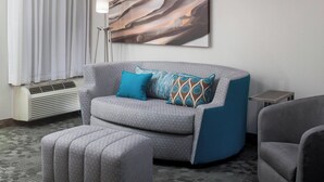 A modern hotel room with a grey sofa, a matching ottoman, a small side table, and a floor lamp. - Courtyard Tuscaloosa by Marriott (Tuscaloosa)