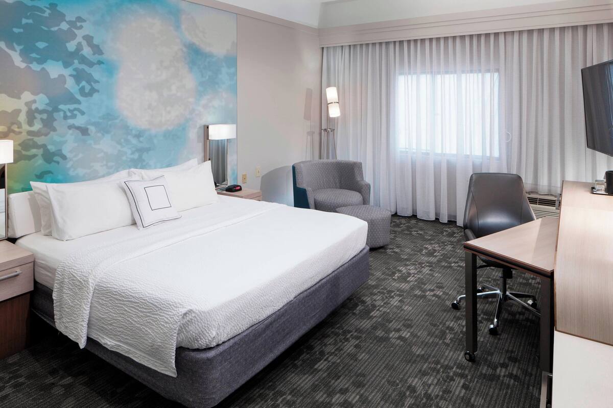 A hotel room with a large bed, a desk, a chair, a flat-screen TV, and a large window with curtains.