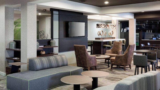 A modern lounge area with sofas, armchairs, and round tables, featuring a flat-screen TV and a decorative wall piece. - Courtyard Tuscaloosa by Marriott (Tuscaloosa)